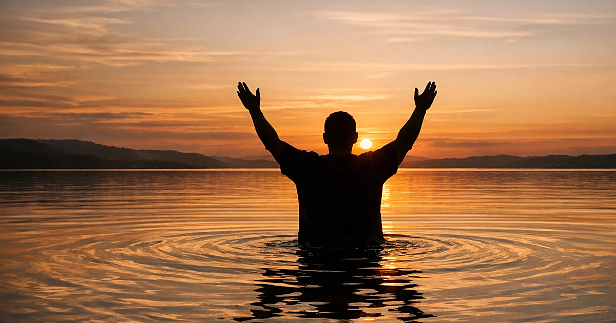 Silhouette of person standing in water at sunset with arms raised upward in spiritual gesture