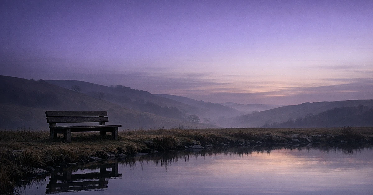 Empty wooden bench beside still pond at twilight with rolling hills in background
