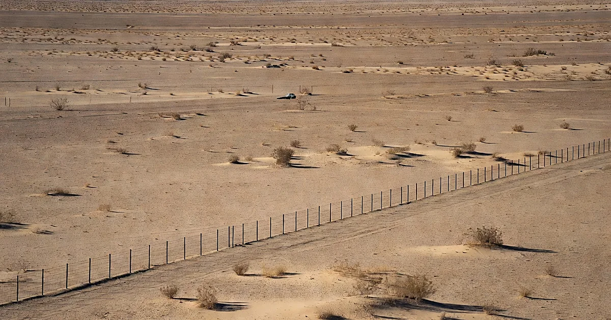 Empty desert border landscape with distant patrol vehicle and metal barrier under bright sunlight